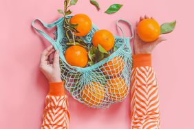 Woman hands in orange striped sweatshirt holding oranges with green leaves in blue eco-friendly shopping mesh bag on pink background.
